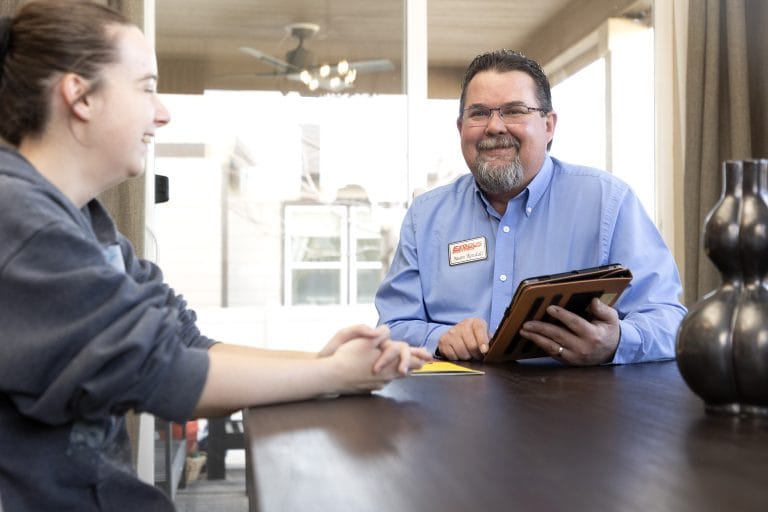 A man with glasses smiling and holding a tablet across from a woman in a bright room.