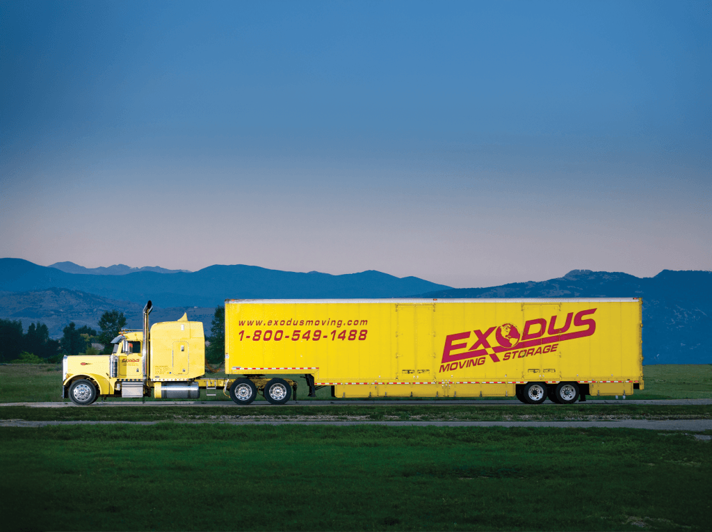 A yellow semi-trailer truck with green fields and mountains in the background.