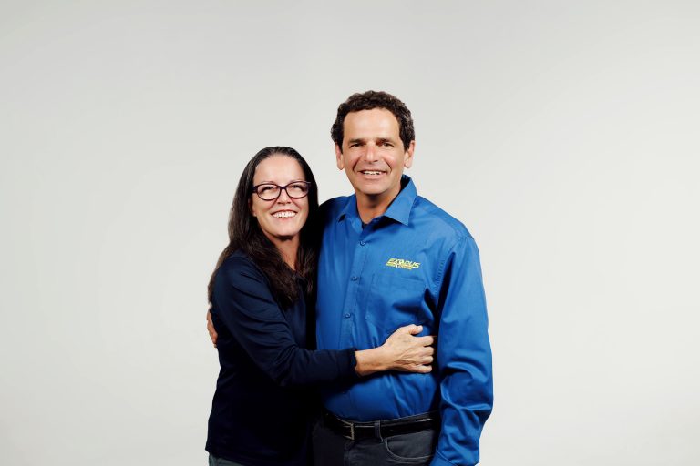 A smiling woman and man standing together in front of a light gray background.