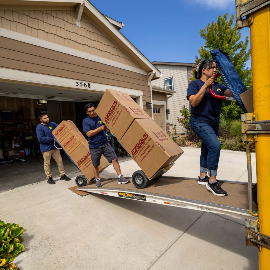 Three people moving cardboard boxes labeled "Exodus Moving" into a truck outside a garage.