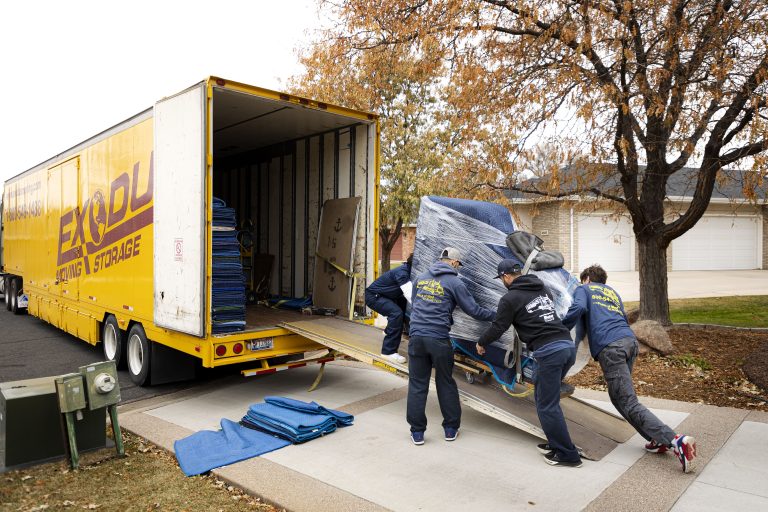 Movers pushing a large wrapped item into a yellow "EXODUS MOVING & STORAGE" truck on a residential street.