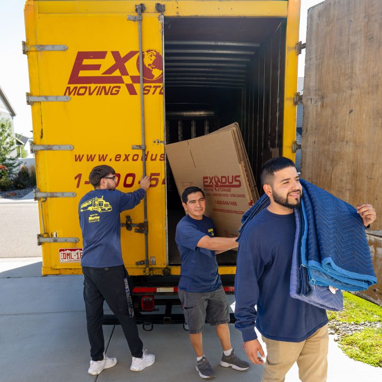 Three people loading a yellow moving truck with a large box.