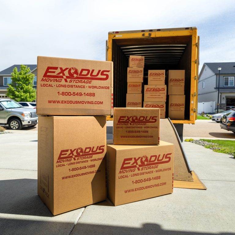 Cardboard boxes labeled "Exodus Moving & Storage" stacked near a yellow truck in a residential area.