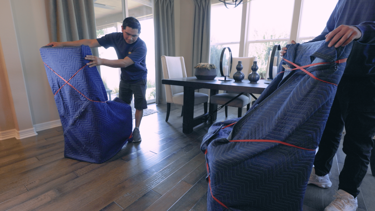 Two individuals moving furniture wrapped in blue quilted blankets secured with red straps in a bright room with wooden floors and a dining set.