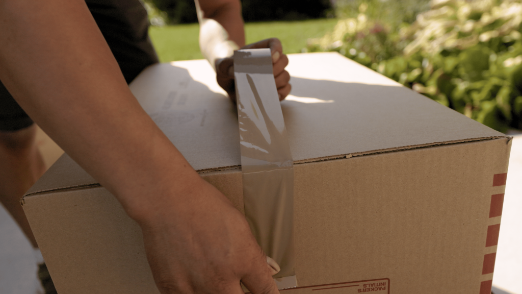 A person sealing a cardboard box with brown packing tape outdoors.