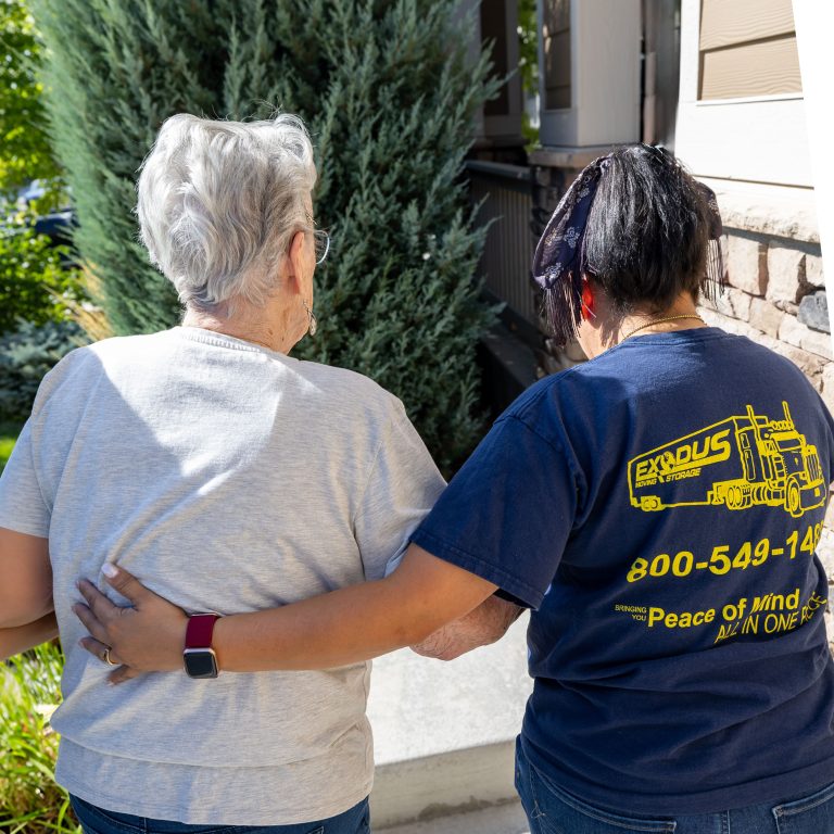 Two women, seen from behind, walk together with arms around each other. One wears a light gray shirt, the other a blue t-shirt with yellow text.