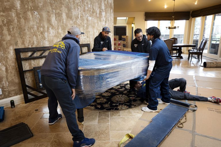 Five people moving a large, wrapped furniture piece in a room with textured walls and a dining area.