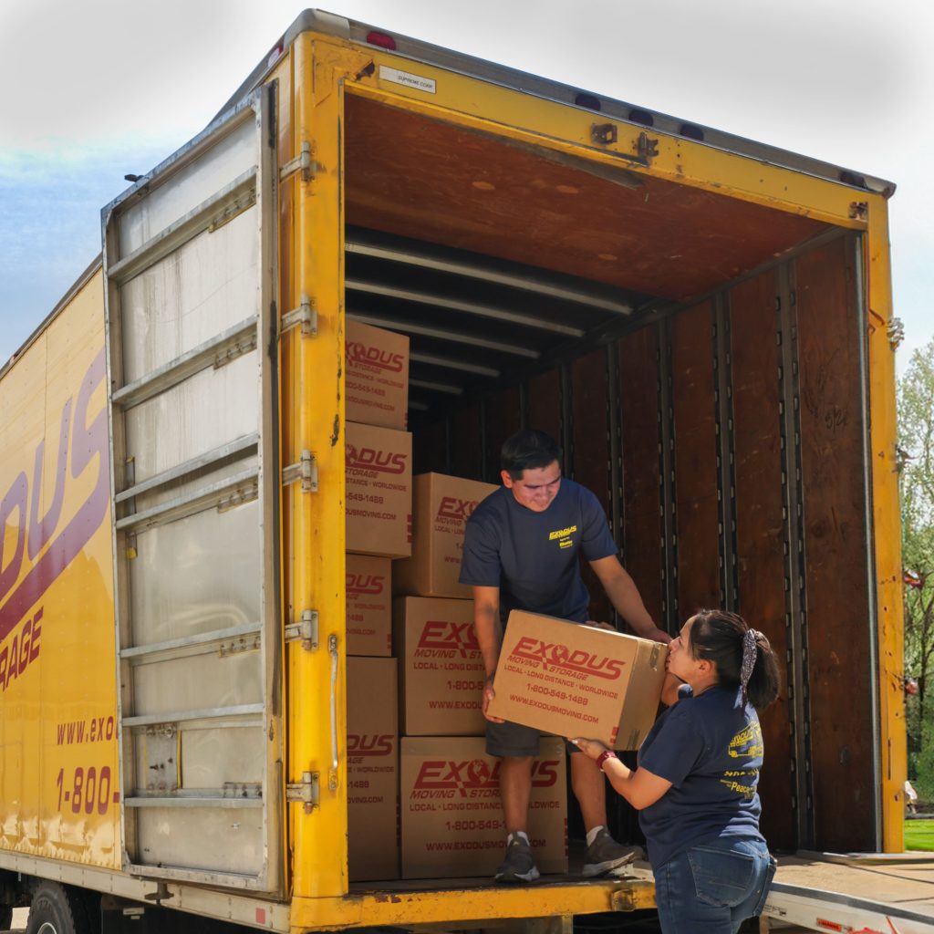 A man and woman loading a moving truck with boxes labeled "EXODUS."